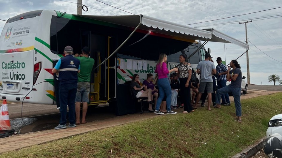 A imagem exibe um ônibus grande, branco e personalizado, estacionado em uma área gramada ao ar livre, servindo como unidade móvel de atendimento.

Ônibus/Unidade Móvel: O veículo está de lado, com a traseira levemente visível à esquerda. Ele é predominantemente branco com detalhes em listras verdes, vermelhas e amarelas. O logo do Governo do Estado do Rio Grande do Sul e a inscrição "Cadastro Único" estão visíveis na lateral. O lado direito do ônibus está totalmente aberto, com uma marquise branca e verde estendida, criando uma área coberta para atendimento.

Pessoas e Atendimento: Um grupo de cerca de dez pessoas (homens e mulheres) está reunido sob a marquise ou em pé próximo à entrada aberta do ônibus. Algumas pessoas estão sentadas em cadeiras dobráveis, enquanto outras conversam em pé. Um homem de coshirt azul e colete de segurança azul (possivelmente um atendente) está de costas para a câmera, próximo à lateral do ônibus.

Ambiente: O ônibus está estacionado em uma área elevada, com um talude gramado em primeiro plano. O fundo mostra uma paisagem de água (possivelmente um rio, lago ou mar, embora não seja claro) e uma palmeira à direita. O céu está nublado, com nuvens cinzentas e brancas. No canto inferior direito, parte de um veículo estacionado está visível.