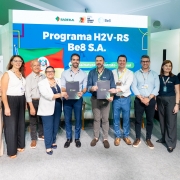 A imagem mostra um grupo de dez pessoas em pé, posando para a foto. Ao centro, o governador Eduardo Leite segura um documento. Ao fundo, se lê "Programa H2V-RS Be8 S.A".
