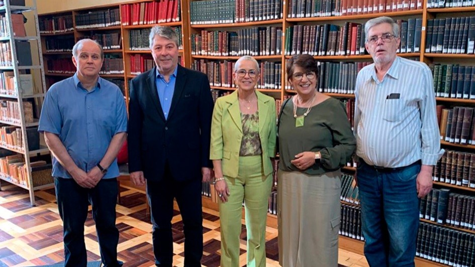 A imagem mostra cinco pessoas em pé, posando para a foto, em uma biblioteca. Ao fundo, há diversas prateleiras cheias de livros catalogados. 