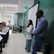 Foto em sala de aula de um homem e uma mulher falando em frente à turma, composta por professores sentados em classes.