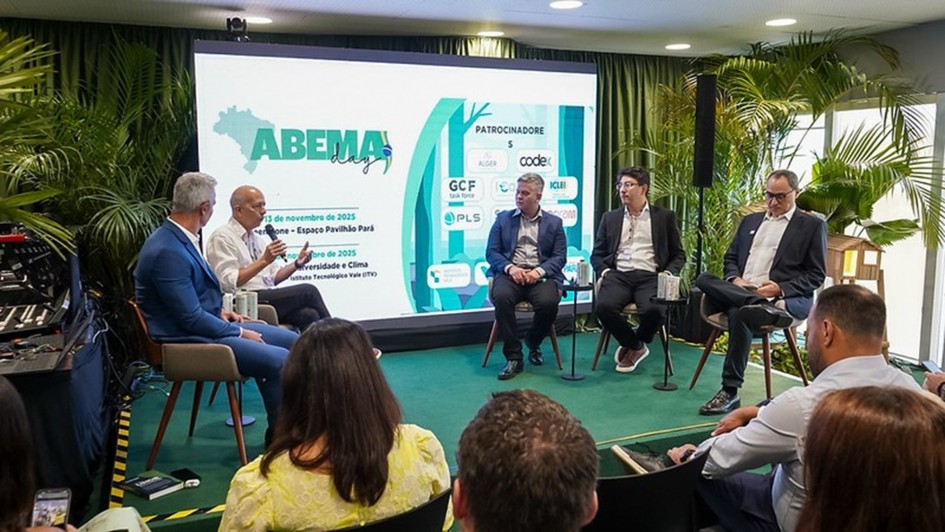 A imagem mostra um painel de debate em um ambiente interno, com decoração em verde e plantas ao fundo. Cinco participantes estão sentados em cadeiras no palco, formando um semicírculo, enquanto conversam diante de uma plateia. Atrás deles, há uma tela grande exibindo o logo “ABEMA Day” e informações sobre datas e patrocínios do evento.