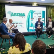 A imagem mostra um painel de debate em um ambiente interno, com decoração em verde e plantas ao fundo. Cinco participantes estão sentados em cadeiras no palco, formando um semicírculo, enquanto conversam diante de uma plateia. Atrás deles, há uma tela grande exibindo o logo “ABEMA Day” e informações sobre datas e patrocínios do evento.