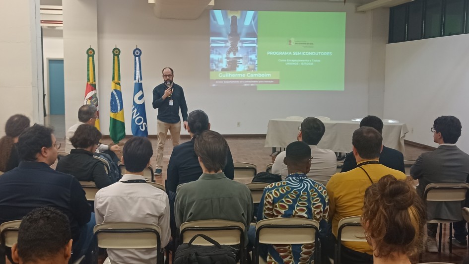 Uma foto em ambiente interno, em uma sala de aula ou auditório. Um homem (Guilherme Cambeim), no centro, está de pé em um estrado de madeira, falando em um microfone que segura na mão direita, voltado para a plateia. Ele veste uma camisa polo escura, calça cáqui e óculos.

Atrás dele, uma tela de projeção exibe um slide com o título "PROGRAMA SEMICONDUTORES", um subtítulo sobre cursos e uma foto vertical com foco em um equipamento ou placa de circuito. O nome "Guilherme Cambeim" aparece em letras menores abaixo.

A plateia é composta por cerca de 20 pessoas (a maioria de costas para a câmera), sentadas em cadeiras brancas em fileiras. No lado esquerdo do estrado, há três mastros com bandeiras: a do Rio Grande do Sul, a do Brasil e uma bandeira com o logotipo da ONU/UNOS. A sala tem paredes brancas e tetos baixos com iluminação de LED embutida.