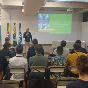 Uma foto em ambiente interno, em uma sala de aula ou auditório. Um homem (Guilherme Cambeim), no centro, está de pé em um estrado de madeira, falando em um microfone que segura na mão direita, voltado para a plateia. Ele veste uma camisa polo escura, calça cáqui e óculos.

Atrás dele, uma tela de projeção exibe um slide com o título "PROGRAMA SEMICONDUTORES", um subtítulo sobre cursos e uma foto vertical com foco em um equipamento ou placa de circuito. O nome "Guilherme Cambeim" aparece em letras menores abaixo.

A plateia é composta por cerca de 20 pessoas (a maioria de costas para a câmera), sentadas em cadeiras brancas em fileiras. No lado esquerdo do estrado, há três mastros com bandeiras: a do Rio Grande do Sul, a do Brasil e uma bandeira com o logotipo da ONU/UNOS. A sala tem paredes brancas e tetos baixos com iluminação de LED embutida.