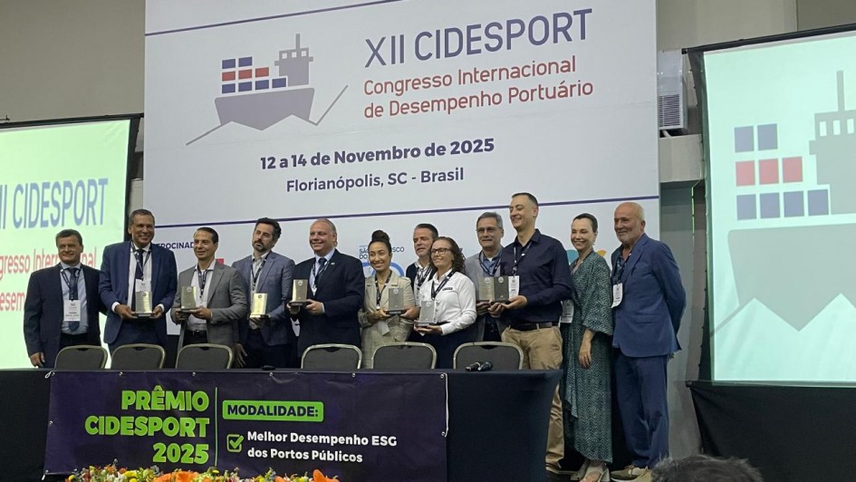 Uma foto tirada em um palco de premiação durante o XII CIDESPORT (Congresso Internacional de Desempenho Portuário). Um grupo de cerca de 10 pessoas (homens e mulheres) está de pé em fila, sorrindo e posando atrás de um pódio forrado com tecido preto. Quase todos estão segurando placas ou troféus retangulares.

Atrás deles, um grande painel de fundo exibe o título "XII CIDESPORT Congresso Internacional de Desempenho Portuário" e as datas "12 a 14 de Novembro de 2025 Florianópolis, SC - Brasil".

Em primeiro plano, sobre a mesa do pódio, há um arranjo de flores em tons de laranja, amarelo e branco. Um cartaz de premiação no pódio diz: "PRÊMIO CIDESPORT 2025" e "MODALIDADE: Melhor Desempenho ESG dos Portos Públicos". A cena é formal, em ambiente de conferência.