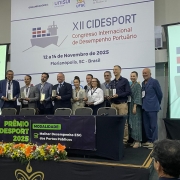 Uma foto tirada em um palco de premiação durante o XII CIDESPORT (Congresso Internacional de Desempenho Portuário). Um grupo de cerca de 10 pessoas (homens e mulheres) está de pé em fila, sorrindo e posando atrás de um pódio forrado com tecido preto. Quase todos estão segurando placas ou troféus retangulares.

Atrás deles, um grande painel de fundo exibe o título "XII CIDESPORT Congresso Internacional de Desempenho Portuário" e as datas "12 a 14 de Novembro de 2025 Florianópolis, SC - Brasil".

Em primeiro plano, sobre a mesa do pódio, há um arranjo de flores em tons de laranja, amarelo e branco. Um cartaz de premiação no pódio diz: "PRÊMIO CIDESPORT 2025" e "MODALIDADE: Melhor Desempenho ESG dos Portos Públicos". A cena é formal, em ambiente de conferência.