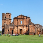 A imagem mostra as ruínas de uma antiga construção missioneira em pedra avermelhada, situadas em um amplo gramado verde. No primeiro plano, um grande crucifixo de madeira aparece desfocado, enquanto ao fundo um grupo de visitantes caminha pelo sítio histórico. O céu está limpo e azul, compondo uma cena típica de turismo cultural ao ar livre.