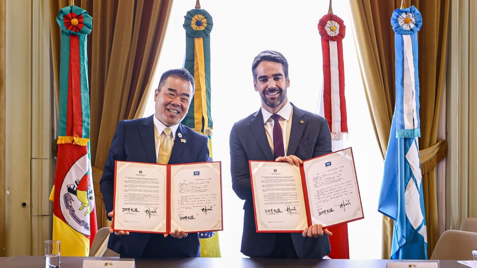 Uma foto de dois homens, o governador do Rio Grande do Sul (à direita, de terno escuro) e o governador da província japonesa de Shiga (à esquerda, de terno e gravata dourada), em pé, posando juntos com cópias abertas de uma declaração conjunta. Ambos sorriem para a câmera e seguram os documentos com as mãos nas laterais e no centro.

A cópia do documento que cada um segura é idêntica, apresentando um texto em inglês no lado esquerdo e em japonês (ou português, dependendo do lado) no lado direito, emoldurado por uma borda vermelha.

Atrás deles, há uma janela central com cortinas grossas de veludo marrom emoldurando-a. Nos cantos, estão hastes de bandeiras ornamentais com fitas coloridas (vermelho, verde e azul) e emblemas redondos: à esquerda, as cores e o brasão do Rio Grande do Sul e, à direita, uma bandeira de cor azul claro e branco.

A mesa em primeiro plano possui placas de identificação com os nomes dos líderes: "TAZO MIKAZUKI" e "EDUARDO LEITE". A cena é formal, em ambiente de reunião de alto nível governamental.