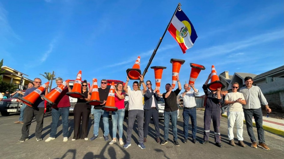 Um grupo de pessoas está alinhado sobre uma via pavimentada, cada uma segurando um cone de trânsito laranja. Uma bandeira grande com as cores azul, vermelha, branca e amarela está hasteada por um dos integrantes. O céu está limpo e azul, e há edificações residenciais ao fundo. Todos estão posicionados lado a lado, com os cones erguidos.