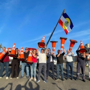 Um grupo de pessoas está alinhado sobre uma via pavimentada, cada uma segurando um cone de trânsito laranja. Uma bandeira grande com as cores azul, vermelha, branca e amarela está hasteada por um dos integrantes. O céu está limpo e azul, e há edificações residenciais ao fundo. Todos estão posicionados lado a lado, com os cones erguidos.