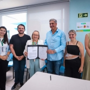 Uma foto em close-up de uma cerimônia de assinatura de convênio em Campo Bom. Seis pessoas (quatro mulheres e dois homens) estão alinhadas, sorrindo e posando atrás de uma mesa branca.

No centro, uma mulher mais velha, de cabelo grisalho e blazer branco sobre calças azul-claro, e um homem alto, de cabelos grisalhos e camisa social azul-clara (manga comprida) e jeans, estão segurando uma pasta preta aberta contendo o documento do convênio. O homem e a mulher segurando a pasta parecem ser as autoridades principais no centro do enquadramento.

À esquerda, duas mulheres vestem blazers azul-claro e escuro, sendo que a primeira usa um crachá pendurado. À direita, uma mulher de blusa preta e saia/calça escura e, ao lado dela, outra mulher de vestido ou casaco longo bege completam o grupo.

A sala tem paredes de cor clara (azul-bebê e branco). No fundo, há uma janela grande e esquadrias azuis que dão para uma área externa. Na parede do fundo, há sinais de segurança ("Saída de Emergência" com seta verde) e um cartaz colorido de aviso ou informação, com o logotipo do governo do Rio Grande do Sul.