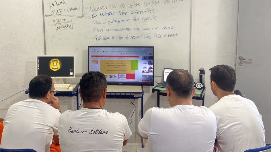 Foto de quatro presidiários sentados de costas para a câmera, acompanhando a transmissão pelo computador numa sala de aula.