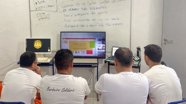 Foto de quatro presidiários sentados de costas para a câmera, acompanhando a transmissão pelo computador numa sala de aula.