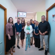 A imagem mostra um grupo de pessoas em pé, posando para a foto em um corredor de um centro médico. A secretária da Saúde está ao centro. 