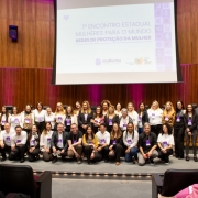 Um grande grupo de participantes posa para uma foto coletiva no auditório, em frente ao palco onde é projetado o título do evento “1º Encontro Estadual Mulheres para o Mundo – Redes de Proteção da Mulher”. As pessoas estão alinhadas em várias fileiras, sorrindo para a câmera.