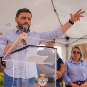 Homem fala ao microfone em evento oficial, atrás de púlpito transparente com brasão. Pessoas acompanham sob tenda, e há mensagem de boas-vindas ao Rio Grande do Sul. Cena pública e institucional.