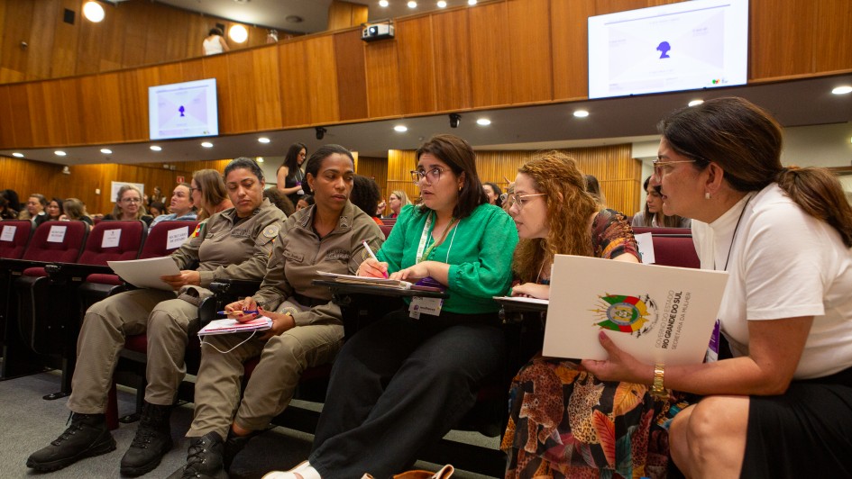 Pessoas sentadas em auditório participam de reunião formal. Alguns usam uniforme, outros traje social. Ambiente com painéis de madeira e telas de apresentação.