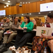 Pessoas sentadas em auditório participam de reunião formal. Alguns usam uniforme, outros traje social. Ambiente com painéis de madeira e telas de apresentação.