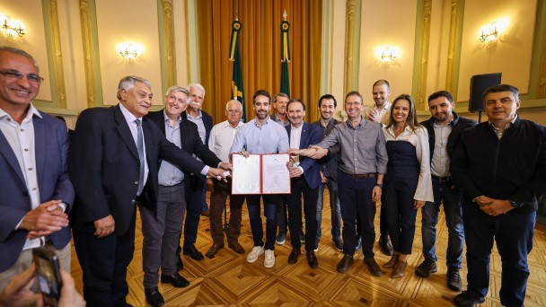 Uma foto de um grupo de treze pessoas (doze homens e uma mulher) em um salão formal, celebrando a autorização de obras. O grupo está posicionado em arco, sorrindo para a câmera, com o governador Eduardo Leite no centro.

Eduardo Leite (de camisa social azul-clara, calças claras e tênis brancos) está segurando um documento aberto (possivelmente a autorização ou convênio) com outros seis homens que também estendem as mãos para tocar a pasta aberta. Os homens vestem camisas sociais, ternos ou blazers. A única mulher no grupo está à direita, vestindo uma calça escura e uma blusa branca com ombros estruturados.

O ambiente é luxuoso, com um piso de madeira em parquet com padrão geométrico de losangos. O fundo tem paredes verde-claras com detalhes em moldura dourada e, no centro, duas bandeiras (a do Brasil e a do Rio Grande do Sul) em mastros altos, emolduradas por cortinas douradas. Em primeiro plano, no canto inferior esquerdo, parte de um celular é visível sendo segurado pela mão de alguém que tira uma foto.