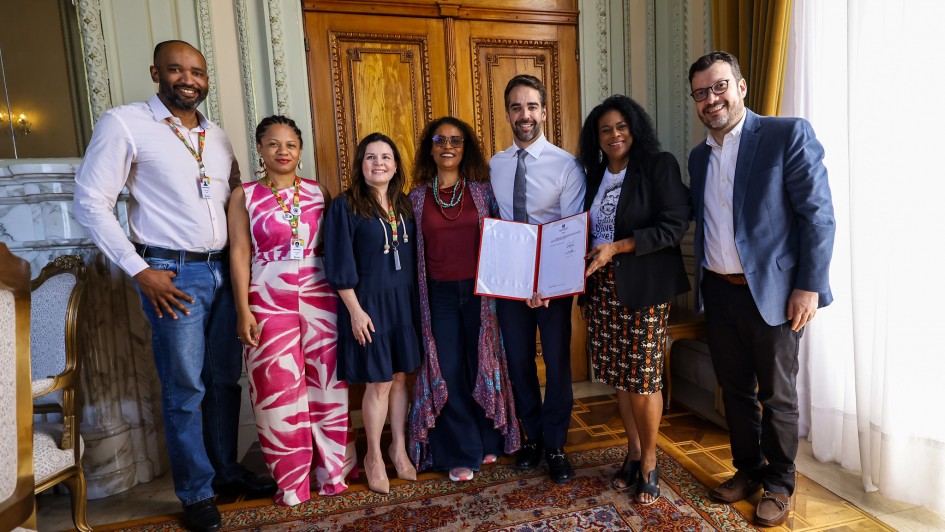 Foto posada de corpo inteiro de sete pessoas de pé em ambiente fechado de decoração clássica. Todos sorriem. Leite está no centro, segurando e exibindo um documento assinado.