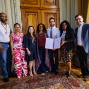 Foto posada de corpo inteiro de sete pessoas de pé em ambiente fechado de decoração clássica. Todos sorriem. Leite está no centro, segurando e exibindo um documento assinado.