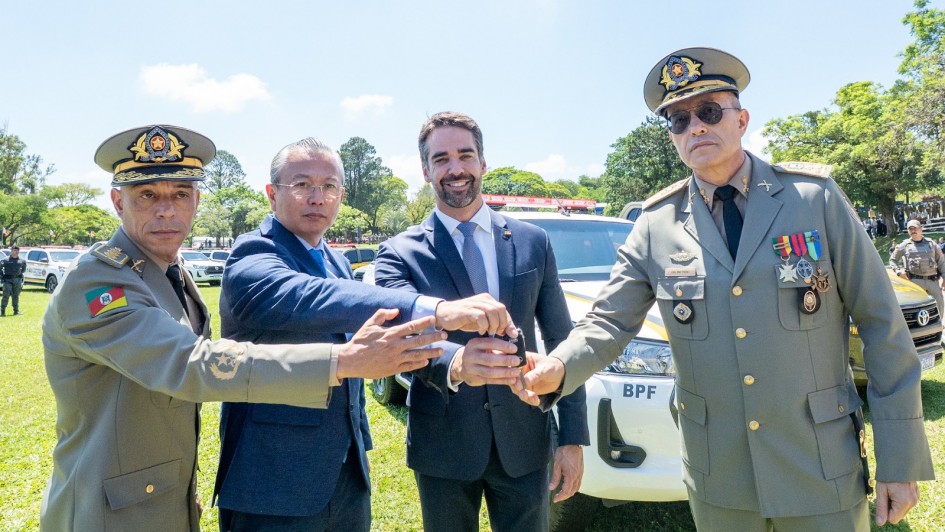 A foto mostra quatro pessoas em pé sobre um gramado, posicionadas em frente a uma viatura policial branca. Todas estão com as mãos estendidas para segurar uma chave. Duas pessoas vestem uniformes militares com insígnias e boinas, enquanto as outras usam roupas sociais. Ao fundo, há mais viaturas alinhadas.