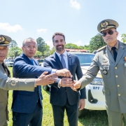A foto mostra quatro pessoas em pé sobre um gramado, posicionadas em frente a uma viatura policial branca. Todas estão com as mãos estendidas para segurar uma chave. Duas pessoas vestem uniformes militares com insígnias e boinas, enquanto as outras usam roupas sociais. Ao fundo, há mais viaturas alinhadas.