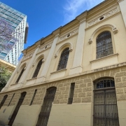 A imagem mostra a fachada de um prédio histórico, com arquitetura clássica e paredes em tom bege. As janelas são altas e arqueadas, protegidas por grades ornamentadas de ferro. A parte inferior da construção apresenta revestimento em pedra trabalhada, enquanto a parte superior exibe ornamentos e detalhes decorativos. À esquerda, parte de uma árvore florida aparece ao lado de um edifício moderno de vidro, criando contraste entre o antigo e o contemporâneo. O céu está azul com poucas nuvens.