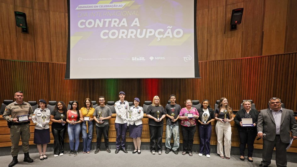 Um grande auditório com painel projetado ao fundo exibindo o texto “Dia Internacional Contra a Corrupção”. Na frente do palco, em linha, estão cerca de 15 pessoas — estudantes, servidores e militares — segurando troféus e placas de premiação. Todos posam para a foto.