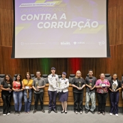 Um grande auditório com painel projetado ao fundo exibindo o texto “Dia Internacional Contra a Corrupção”. Na frente do palco, em linha, estão cerca de 15 pessoas — estudantes, servidores e militares — segurando troféus e placas de premiação. Todos posam para a foto.