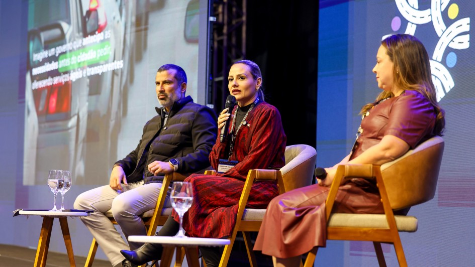 A foto mostra três pessoas sentadas em um palco. A do meio está falando ao microfone. Ao fundo, há um telão colorido. 