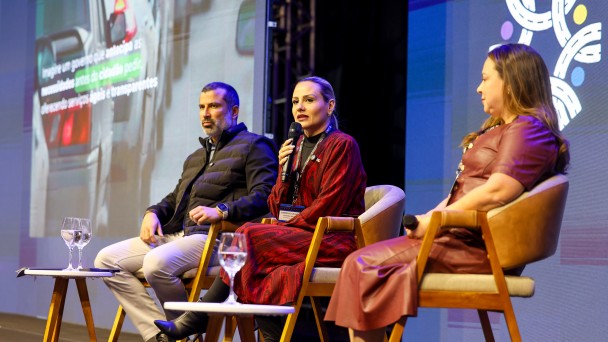 A foto mostra três pessoas sentadas em um palco. A do meio está falando ao microfone. Ao fundo, há um telão colorido. 