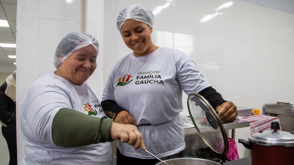 Foto em plano americano de duas mulheres cozinhando em um panela no fogão. Elas estão numa cozinha e usam a camiseta do projeto.