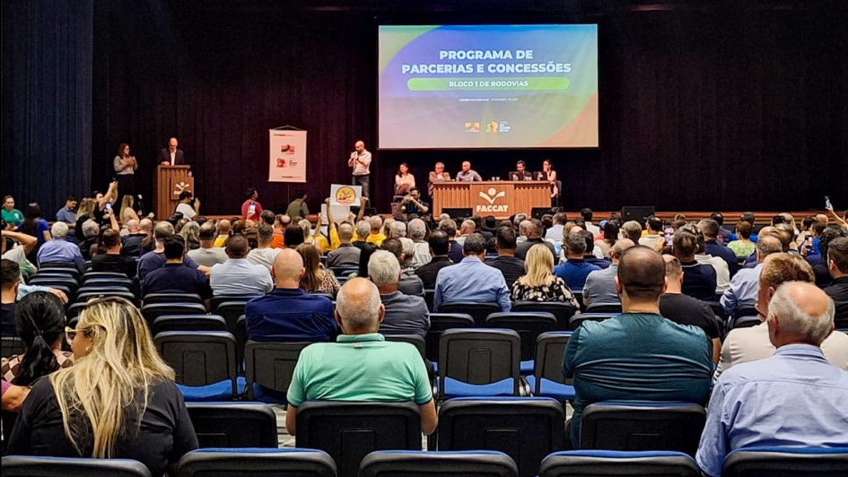 A foto registra uma audiência pública em um auditório grande e lotado, com fileiras de cadeiras ocupadas. No palco, há uma mesa com integrantes da apresentação e um telão exibindo o título “Programa de Parcerias e Concessões – Bloco 1 de Rodovias”. À esquerda, uma pessoa fala ao microfone em um púlpito. O público preenche todo o espaço visível, demonstrando ampla participação no evento.
