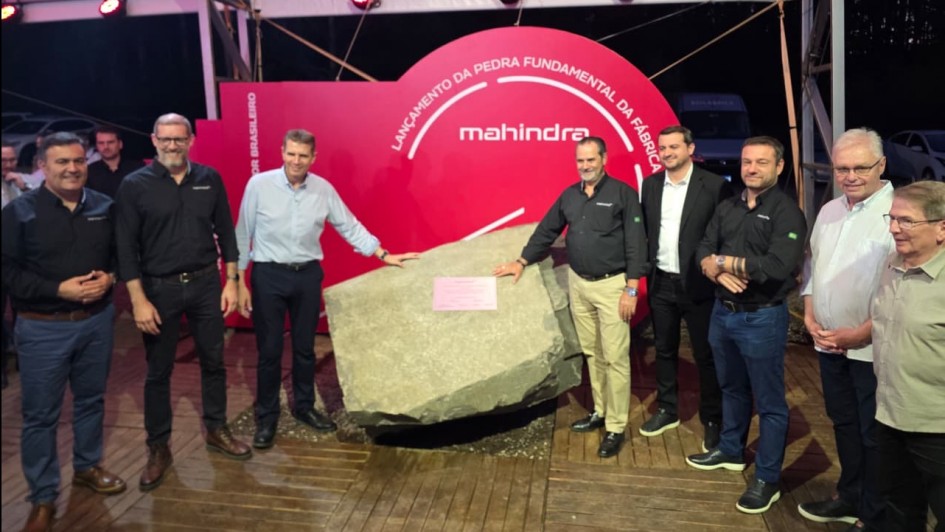 A imagem mostra um grupo de homens posando diante de uma grande pedra instalada sobre uma base de madeira, sob uma estrutura coberta. Atrás deles há um painel vermelho semicircular com o texto “Lançamento da Pedra Fundamental da Fábrica” e a marca “Mahindra”. O grupo está alinhado, olhando para a câmera, alguns com a mão apoiada sobre a pedra.