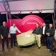 A imagem mostra um grupo de homens posando diante de uma grande pedra instalada sobre uma base de madeira, sob uma estrutura coberta. Atrás deles há um painel vermelho semicircular com o texto “Lançamento da Pedra Fundamental da Fábrica” e a marca “Mahindra”. O grupo está alinhado, olhando para a câmera, alguns com a mão apoiada sobre a pedra.