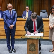 A cena mostra uma cerimônia formal em um auditório. No centro, um homem de terno preto e gravata vermelha assina um documento sobre uma mesa de vidro com um brasão na frente. Ao redor dele, três pessoas em pé observam: um homem de terno azul e duas mulheres, uma segurando uma pasta aberta. Ao fundo, outras pessoas sentadas em uma mesa grande de madeira acompanham o ato. O ambiente é solene, com iluminação suave e painel de madeira atrás.