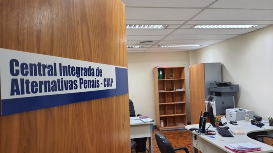 A imagem mostra a porta de uma sala com mesas de escritório, computador, cadeiras e estantes. Na porta, em destaque, se lê "Central Integrada de Alternativas Penais - CIAP". 