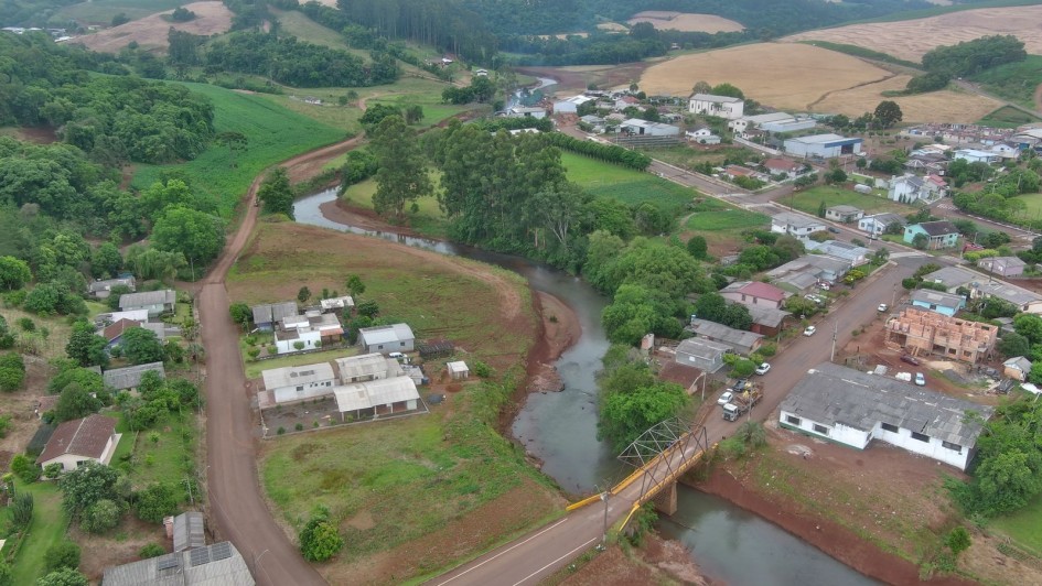 Uma foto aérea de uma área urbana e rural em um vale, cortada por um rio sinuoso. O rio, de águas turvas e leito largo (sugerindo desassoreamento), passa pelo centro da imagem, cercado por vegetação densa e terrenos de cor marrom-avermelhada.

Na margem direita do rio, em primeiro plano, uma pequena ponte de treliça metálica, pintada de amarelo, cruza o rio. Próximo à ponte, há uma rua asfaltada com alguns veículos e casas simples de telhado escuro, algumas com fachadas brancas.

O lado esquerdo do rio mostra um aglomerado de casas mais antigas. No fundo, a paisagem se eleva em colinas cobertas por campos verdes e áreas de plantio de cor amarelo-claro/marrom, com grandes fileiras de árvores em um plano intermediário. O céu é nublado e de cor clara. A foto ilustra a paisagem de uma cidade com foco em um rio sendo recuperado ou manejado.