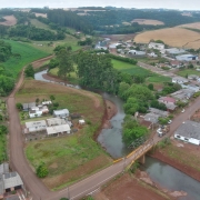Uma foto aérea de uma área urbana e rural em um vale, cortada por um rio sinuoso. O rio, de águas turvas e leito largo (sugerindo desassoreamento), passa pelo centro da imagem, cercado por vegetação densa e terrenos de cor marrom-avermelhada.

Na margem direita do rio, em primeiro plano, uma pequena ponte de treliça metálica, pintada de amarelo, cruza o rio. Próximo à ponte, há uma rua asfaltada com alguns veículos e casas simples de telhado escuro, algumas com fachadas brancas.

O lado esquerdo do rio mostra um aglomerado de casas mais antigas. No fundo, a paisagem se eleva em colinas cobertas por campos verdes e áreas de plantio de cor amarelo-claro/marrom, com grandes fileiras de árvores em um plano intermediário. O céu é nublado e de cor clara. A foto ilustra a paisagem de uma cidade com foco em um rio sendo recuperado ou manejado.
