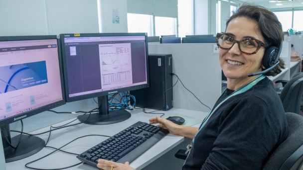 A imagem mostra uma consultora do Telessaúde sentada em frente a sua mesa de trabalho. Ela olha para a câmera e, na sua frente, há dois monitores de computador. 
