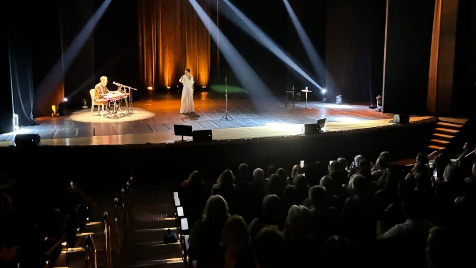 Apresentação ao vivo em teatro com duas pessoas no palco, luzes dramáticas e público atento nas cadeiras. Atmosfera artística e envolvente.