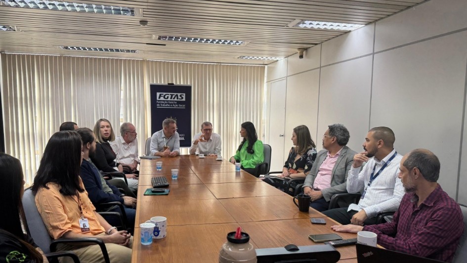 Foto mostra reunião na sala da Fgtas, com pessoas sentadas a volta de uma mesa de trabalho. O banner da Fgtas está atrás dos secretários.