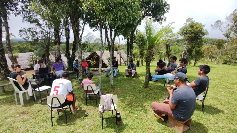 A imagem mostra um grupo de pessoas reunidas em círculo ao ar livre, em uma área gramada e cercada por árvores. Algumas estão sentadas em cadeiras simples, outras em bancos de madeira. Ao fundo, há pequenas construções de madeira e uma paisagem de morros e vegetação densa. O encontro ocorre sob a sombra das árvores, com participantes conversando ou anotando informações. O ambiente remete a uma reunião comunitária em contexto rural ou indígena, em meio à natureza.