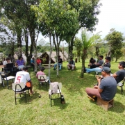 A imagem mostra um grupo de pessoas reunidas em círculo ao ar livre, em uma área gramada e cercada por árvores. Algumas estão sentadas em cadeiras simples, outras em bancos de madeira. Ao fundo, há pequenas construções de madeira e uma paisagem de morros e vegetação densa. O encontro ocorre sob a sombra das árvores, com participantes conversando ou anotando informações. O ambiente remete a uma reunião comunitária em contexto rural ou indígena, em meio à natureza.
