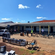 A imagem mostra o pátio externo de uma escola, com diversas cadeiras e equipamentos organizados ao ar livre. Algumas pessoas caminham e inspecionam o local. O prédio escolar ao fundo apresenta estrutura simples, com telhado de duas águas e portas distribuídas ao longo da fachada. O céu está limpo e ensolarado.