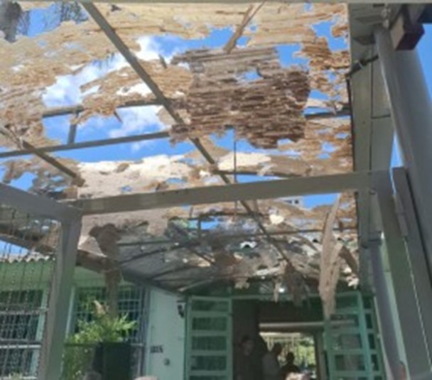 A imagem mostra a cobertura externa danificada de uma escola. O telhado translúcido está completamente perfurado, com grandes áreas destruídas, expondo o céu. Debaixo da estrutura, parte da entrada da escola é visível.
