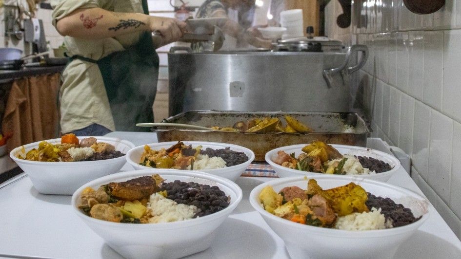 A imagem mostra em primeiro plano cinco marmitas em embalagens plásticas, contendo arroz, feijão, carne e legumes. Ao fundo, há duas pessoas cozinhando, mexendo em panelas grandes no fogão. 