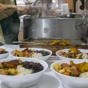 A imagem mostra em primeiro plano cinco marmitas em embalagens plásticas, contendo arroz, feijão, carne e legumes. Ao fundo, há duas pessoas cozinhando, mexendo em panelas grandes no fogão. 