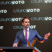 Um homem, presumivelmente o Governador Eduardo Leite, está em pé atrás de um púlpito de cor escura, fazendo um discurso. Ele veste um terno azul-marinho, uma camisa branca e uma gravata roxa escura. Ele tem barba e cabelo curto e escuro. A mão direita dele está apoiada sobre o púlpito, e a mão esquerda está levantada ao lado, com a palma aberta, como se estivesse gesticulando enquanto fala. O fundo é escuro, e as palavras "GRUPO VOTO" estão visíveis repetidamente em letras maiúsculas brancas atrás e acima dele. Um grande 'V' estilizado em dourado ou laranja é visível na parte frontal do púlpito. A iluminação está focada no orador, tornando o fundo menos distinto.